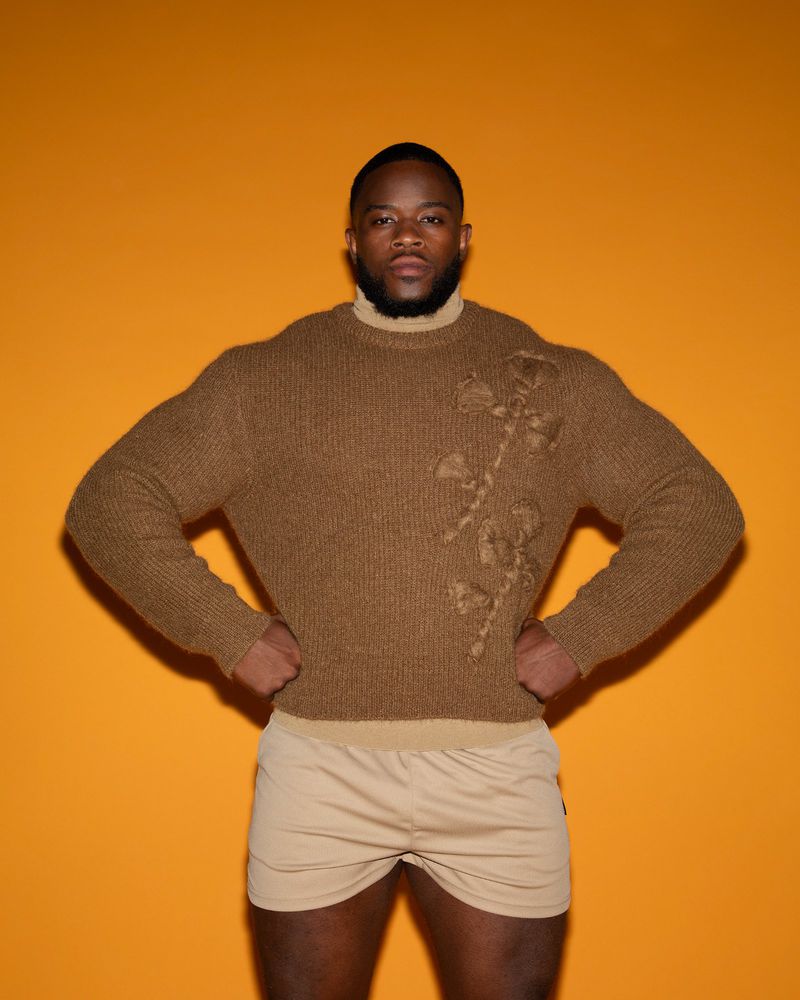 Adult man in brown floral-detailed sweater posing indoors against an orange background with hands on hips, wearing beige shorts.