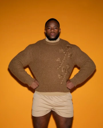 Adult man in brown floral-detailed sweater posing indoors against an orange background with hands on hips, wearing beige shorts.