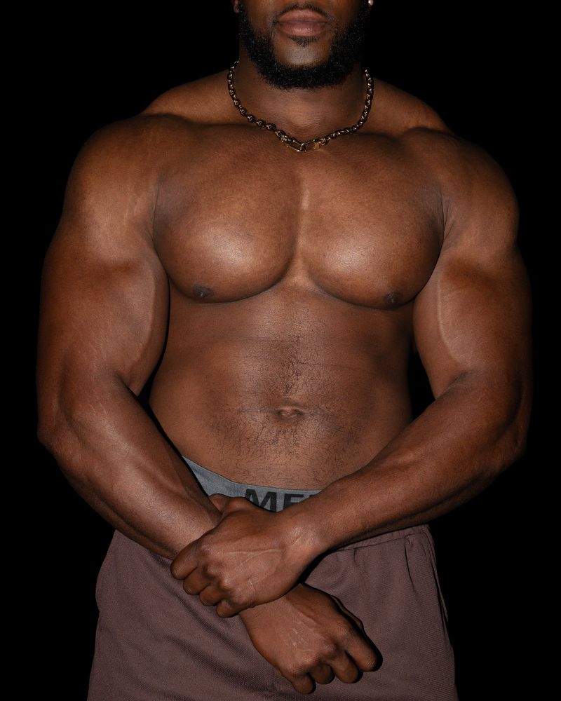 An adult African-American male bodybuilder poses shirtless with arms crossed, showcasing muscular physique against a dark backdrop.