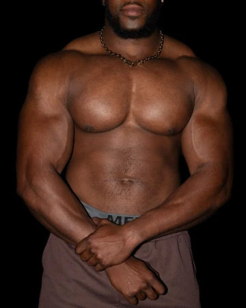 An adult African-American male bodybuilder poses shirtless with arms crossed, showcasing muscular physique against a dark backdrop.