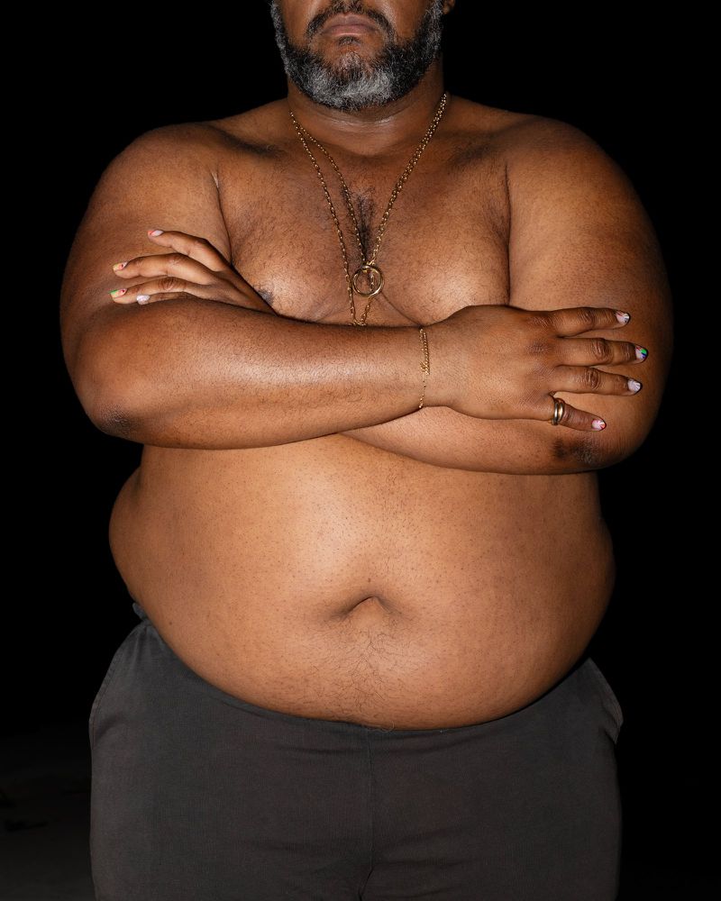 Upper torso shot of a dark-skinned adult male with arms crossed, dark hair, and a graying beard, set against a black backdrop.