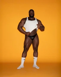Muscular Black man posing in a white tank top, black briefs, and white socks against a bright yellow seamless backdrop.