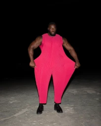 Muscular Black man wearing a wide pleated hot pink jumpsuit and black loafers against a dark backdrop.
