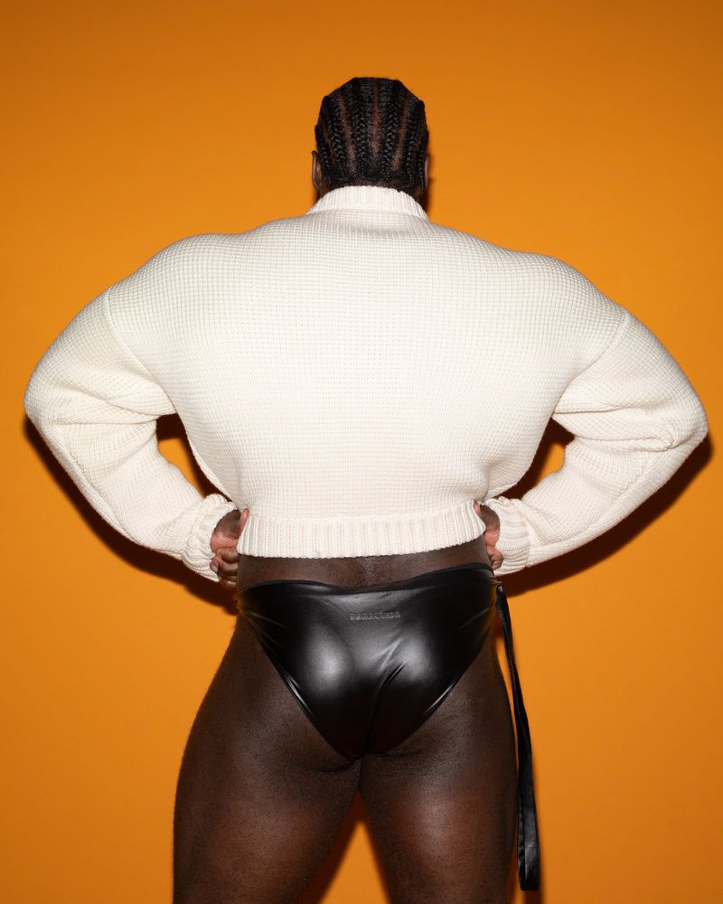 Rear view of a Black man with braided hair wearing a white textured sweater and black briefs against an orange background.