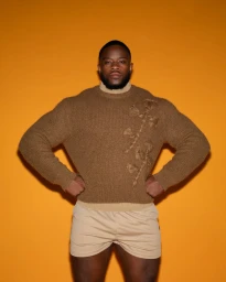 Black man standing against a yellow background wearing a brown textured sweater and beige shorts
