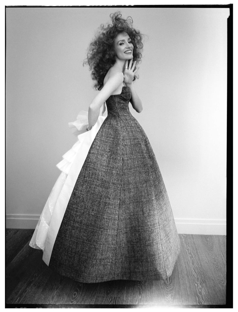 Black and white portrait of a smiling adult woman in a strapless gown with ruffled detail posing in front of a plain wall.
