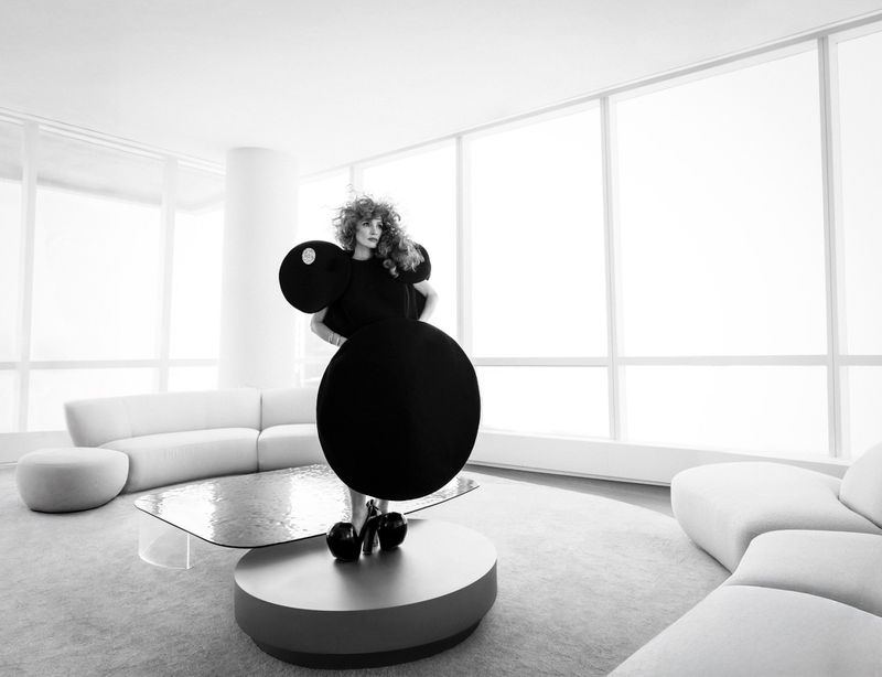 A woman with curly hair poses in a black, sculptural dress with circular shapes in a modern, bright living room interior.