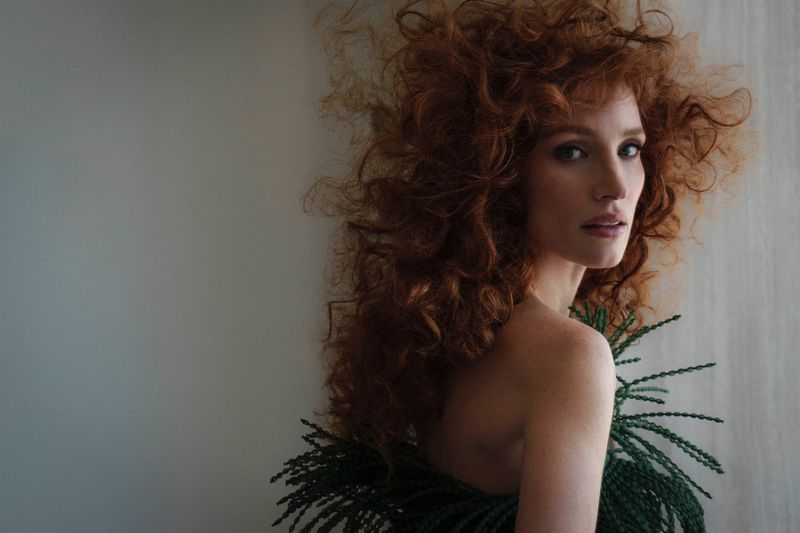 Portrait of a red-haired woman with elaborate curls wearing green foliage around her neck, looking over her bare shoulder at the camera.