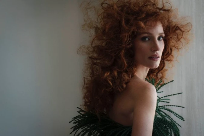 Portrait of a red-haired woman with elaborate curls wearing green foliage around her neck, looking over her bare shoulder at the camera.