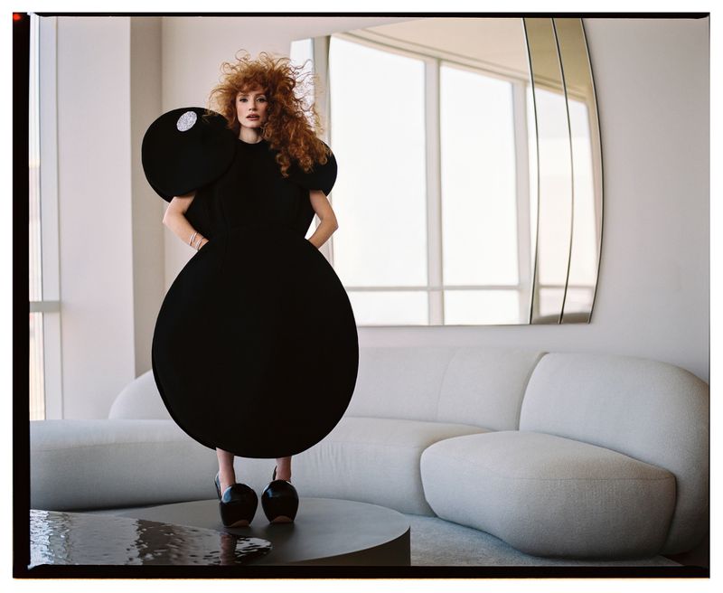 A woman with red hair poses in a dark, round, avant-garde dress in a bright indoor setting with a modern white couch and mirror.