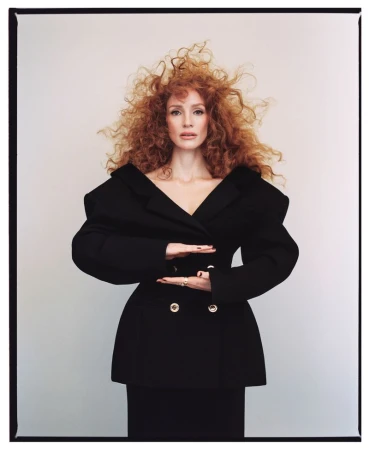 Jessica Chastain poses in a black blazer and skirt with red curly hair against a neutral background, with a medium shot framing her.