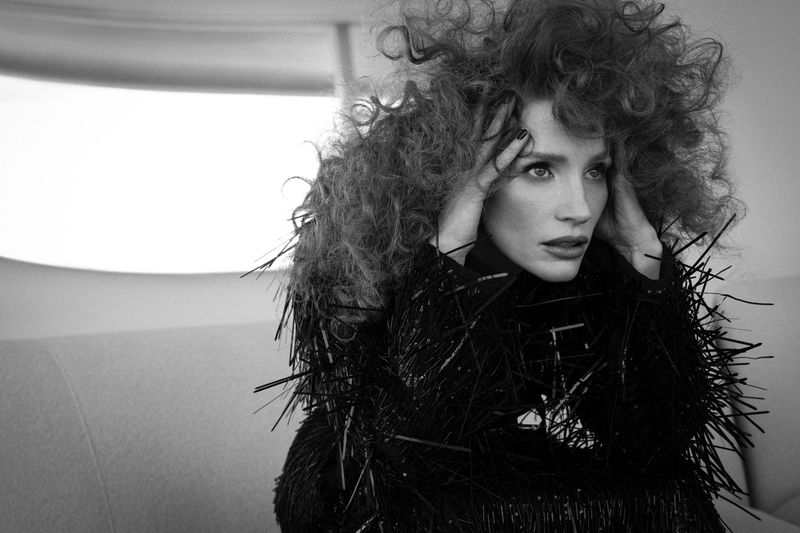 Black and white portrait of Jessica Chastain with hands in her curly red hair, wearing a spiky black top, in a studio environment.