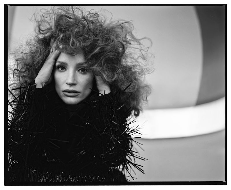 Black and white portrait of a fair-skinned woman with curly hair and a fringed top, looking at the camera with hands near her face.