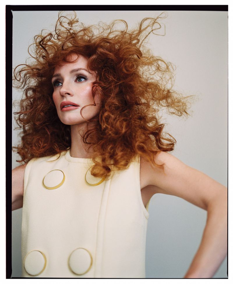 A woman with curly red hair poses in a sleeveless dress with circular buttons against a neutral background in a fashion portrait.