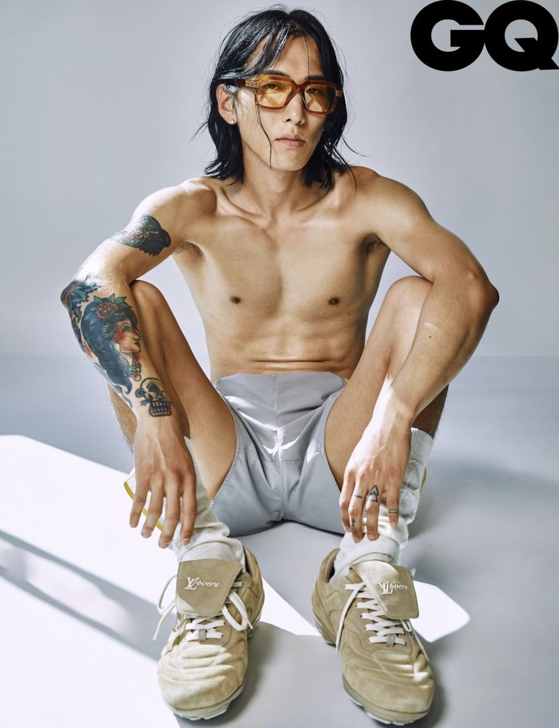 Shirtless person sits in tan sneakers, light grey shorts & sunglasses. Tattooed man with long dark hair poses on plain white background.