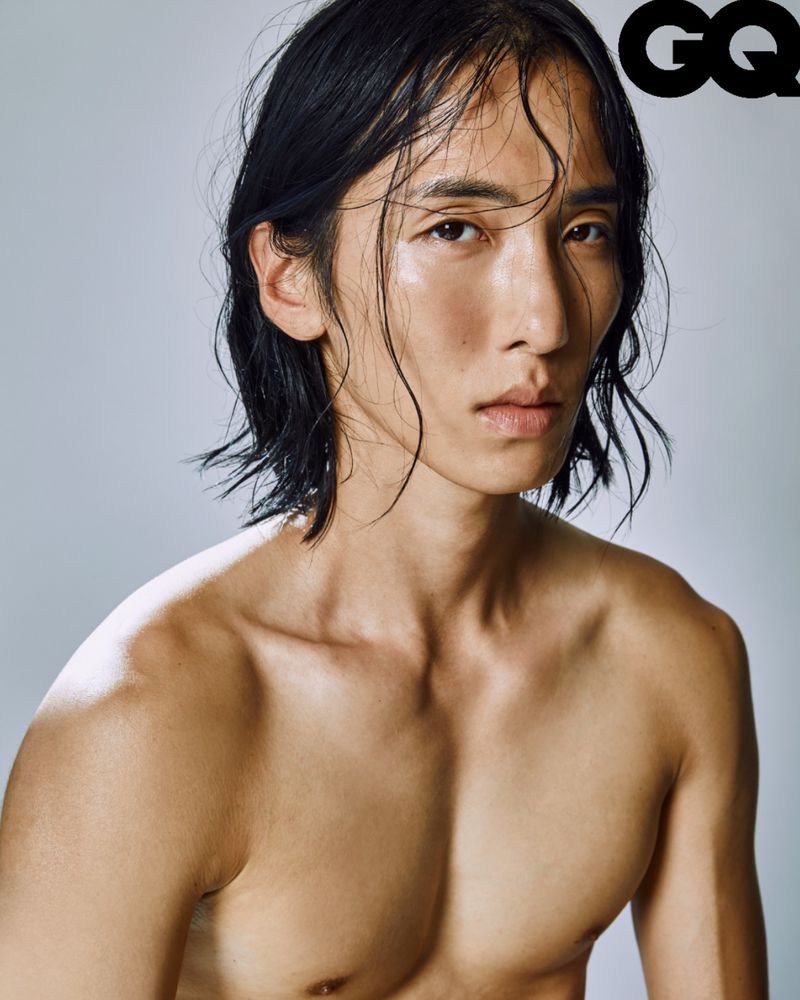 Headshot of an adult male with long black hair, bare shoulders, and light skin, set against a neutral background with the GQ logo.