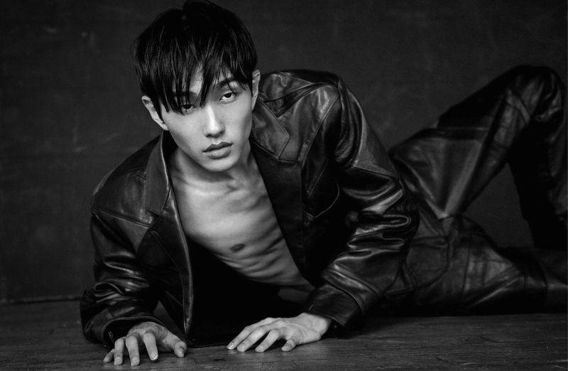 Portrait of a shirtless adult male with black hair wearing a black leather jacket, posed reclining on the floor in a photo studio, black and white.