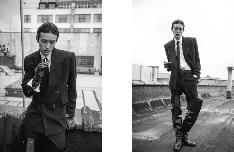 Monochrome fashion shot of an Asian male model in a suit and boots on a rooftop against an urban building backdrop.