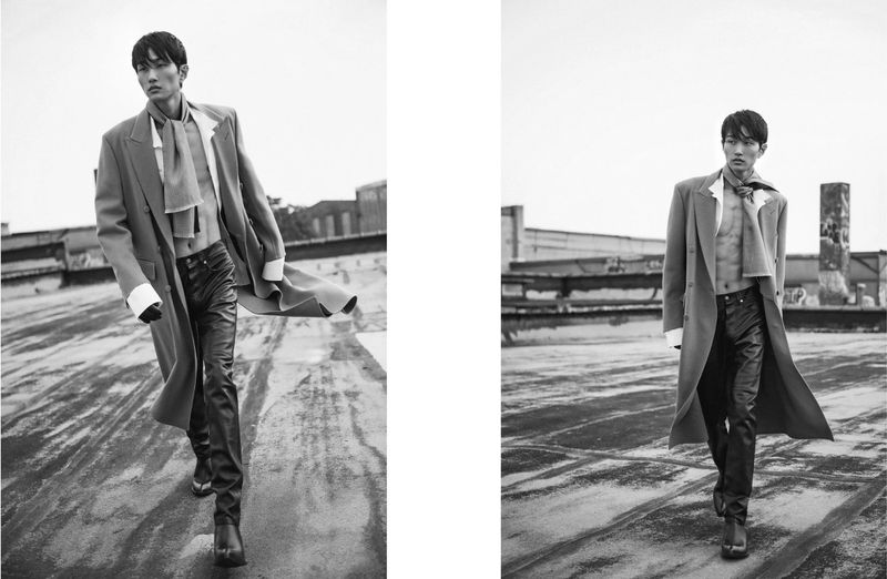 Black and white fashion portrait of a slender Asian male model in a long coat, dress shirt, scarf, and leather pants on a rooftop.