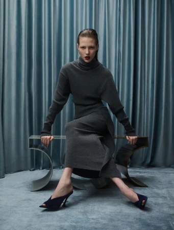 Fashion shot of a woman in a grey turtleneck dress and blue high-heeled shoes, sitting on a glass bench with a blue draped background.