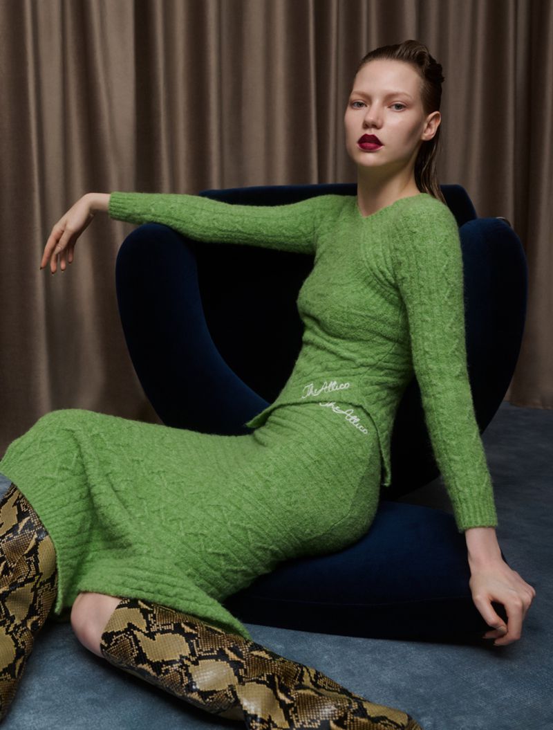 A fashionable woman with dark red lipstick poses while sitting in a chair, wearing a matching green knitted sweater and skirt with snakeskin boots.