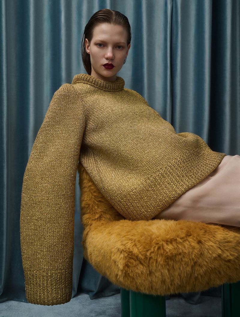 Fashion shot of a woman in a gold knit turtleneck sweater sitting on a green-legged chair with a fuzzy gold cushion against a teal curtain.