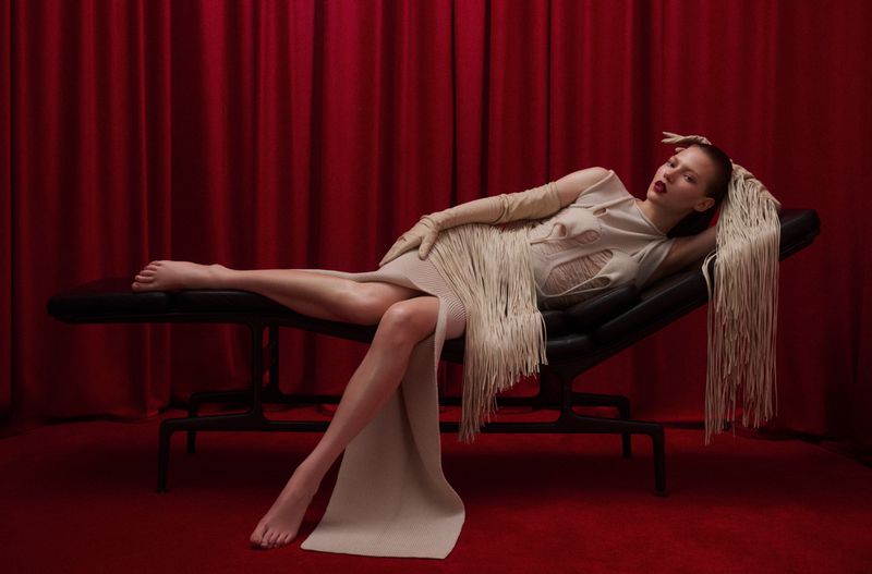 Adult woman in fringed beige dress reclines on black chaise lounge, with red carpet and curtain backdrop in a fashion editorial.