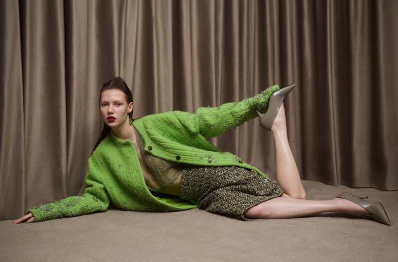 A fashion shot of a woman in a green knit cardigan and neutral heels, lying on her side against a beige curtain backdrop.