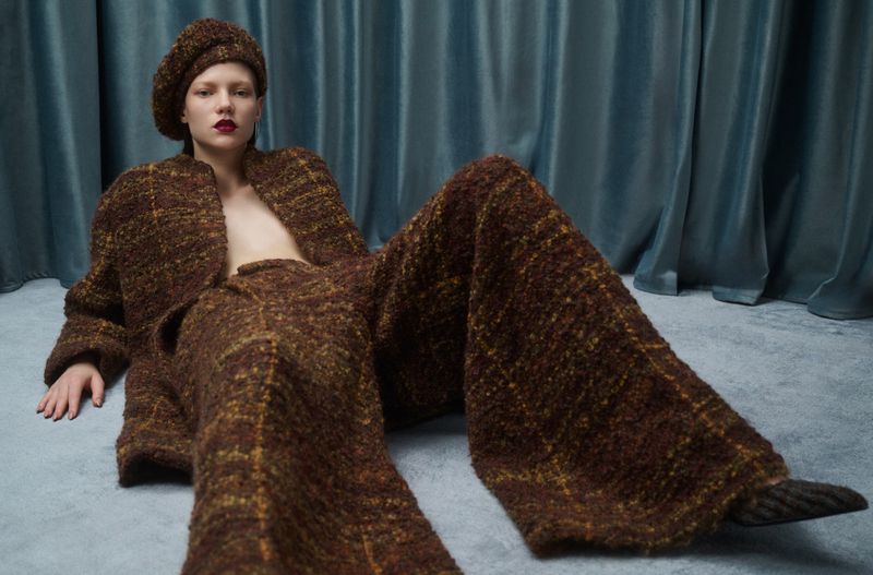 A fair-skinned adult female model is seated on a gray carpet wearing a brown and yellow tweed matching jacket, wide-leg pants, and beret.