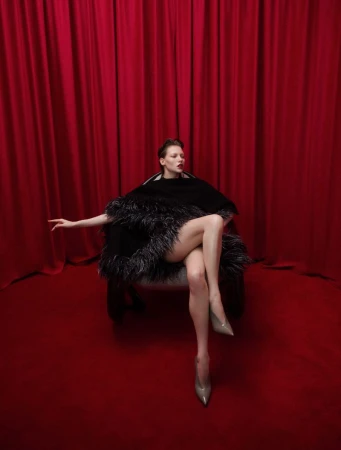 Elegant woman with dark hair poses in a feathered cape and high heels against a red backdrop. Fashion, style, and glamour.
