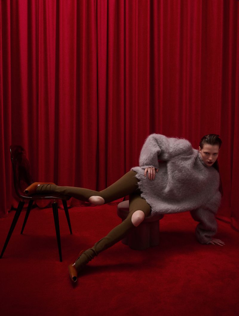 A person in a grey sweater and green leggings poses on a red carpet in front of a red curtain, with a black chair to the left.