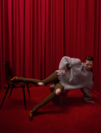 A person in a grey sweater and green leggings poses on a red carpet in front of a red curtain, with a black chair to the left.