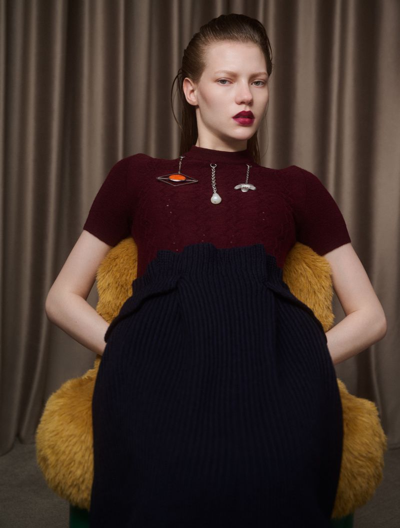 Adult female in studio wearing maroon top and dark skirt sitting in furry yellow chair with neutral curtain background.