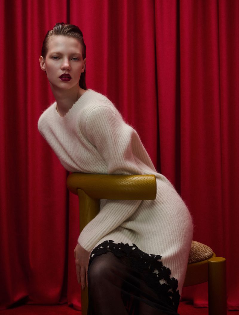 Fashion image of a woman in a white sweater and black skirt sitting on a yellow chair in front of a red curtain backdrop.