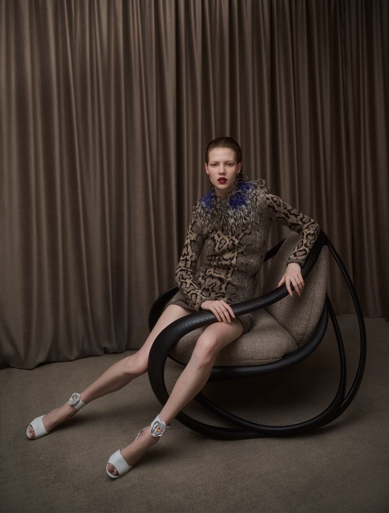 A poised fashion model sits in a black rocking chair, wearing a patterned dress and white heels in a photo studio setting.
