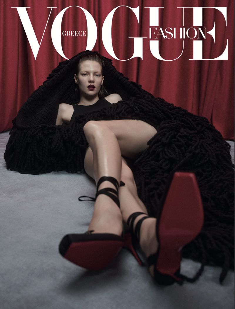 Vogue Greece fashion magazine cover featuring a fashion model in black shoes and a black outfit with a red backdrop.