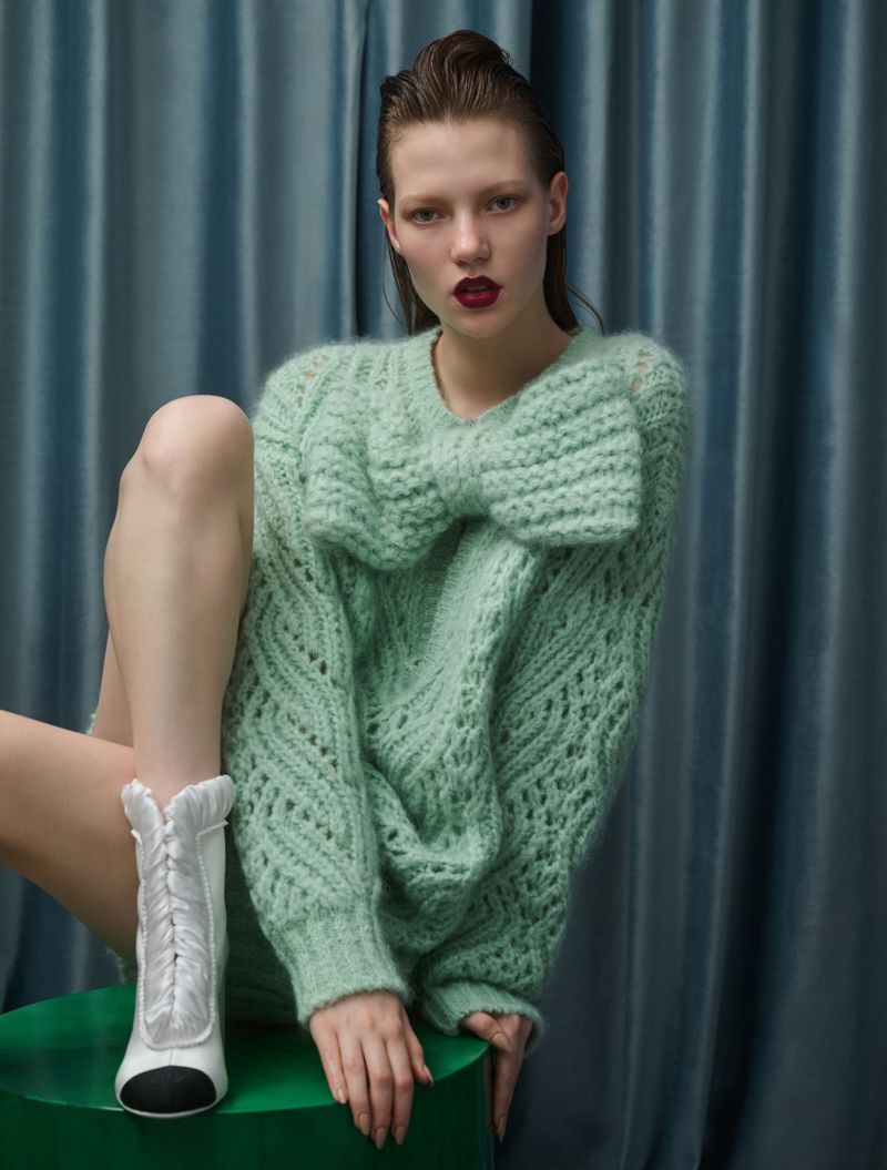 Fashion editorial shot of a young adult female model with dark lipstick in a mint green sweater with a bow, and white ankle boots.