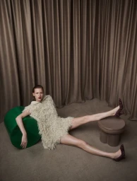 Model wearing a textured fringed dress reclining against a green cylindrical prop with a brown stool