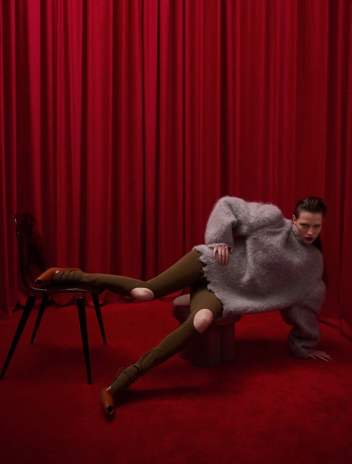 Model in fluffy grey sweater and brown tights posing against deep red curtains for Vogue Greece