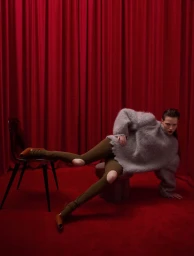 Model in fluffy grey sweater and brown tights posing against deep red curtains for Vogue Greece
