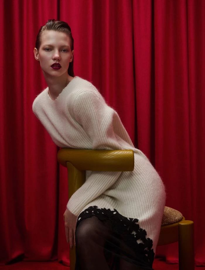 Model in white knit dress leaning over a yellow chair against pleated red curtains