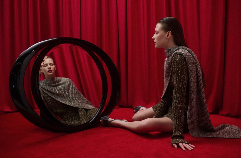 Model seated on a red carpet in front of red drapes next to a large circular black mirror reflecting her image.