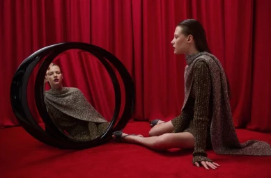 Model seated on a red carpet in front of red drapes next to a large circular black mirror reflecting her image.