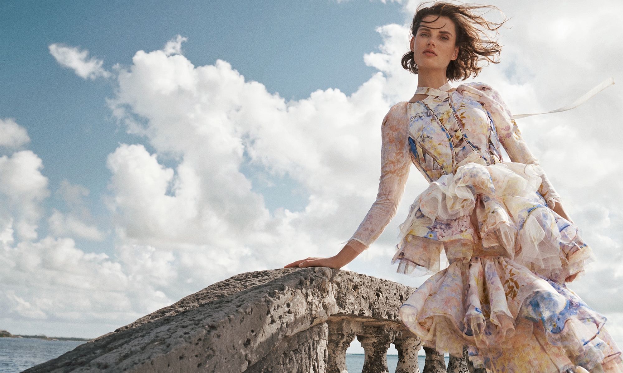 Woman in an elaborate floral dress stands on a stone structure against a sky with puffy clouds, fashion editorial shot.