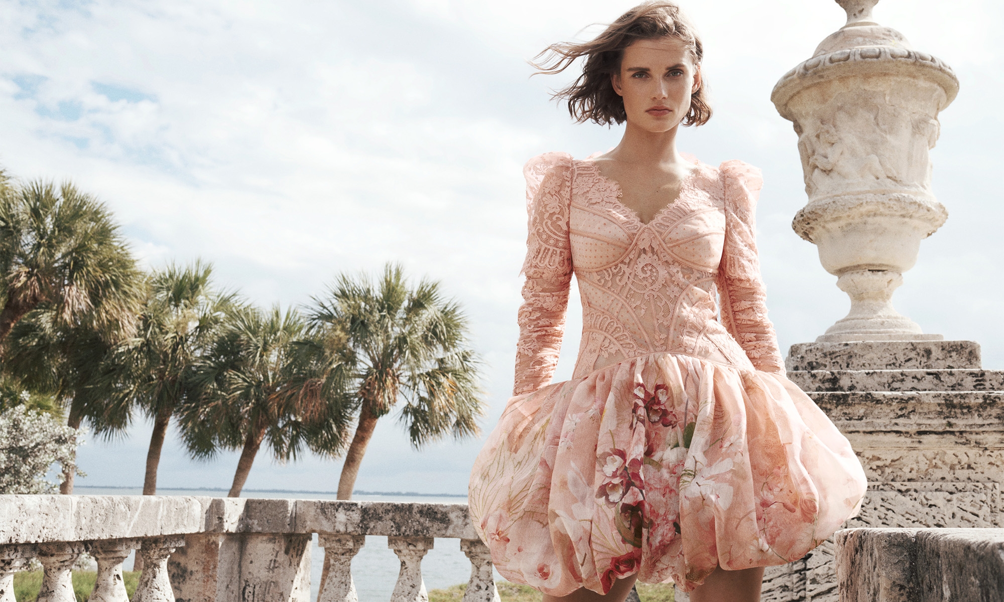 Woman in pink floral gown poses on ornate balcony overlooking ocean with palm trees and cloudy sky in a fashion or beauty editorial shot.