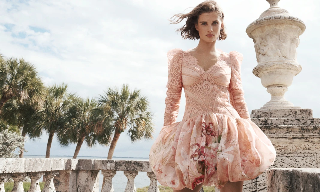 Woman in pink floral gown poses on ornate balcony overlooking ocean with palm trees and cloudy sky in a fashion or beauty editorial shot.