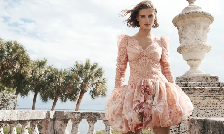 Woman in pink floral gown poses on ornate balcony overlooking ocean with palm trees and cloudy sky in a fashion or beauty editorial shot.