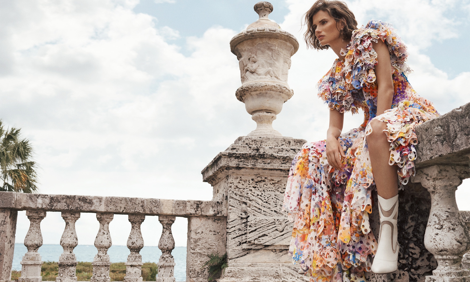 A woman in a floral dress and white boots sits on a stone balustrade with a decorative urn in the background, overlooking the sea.