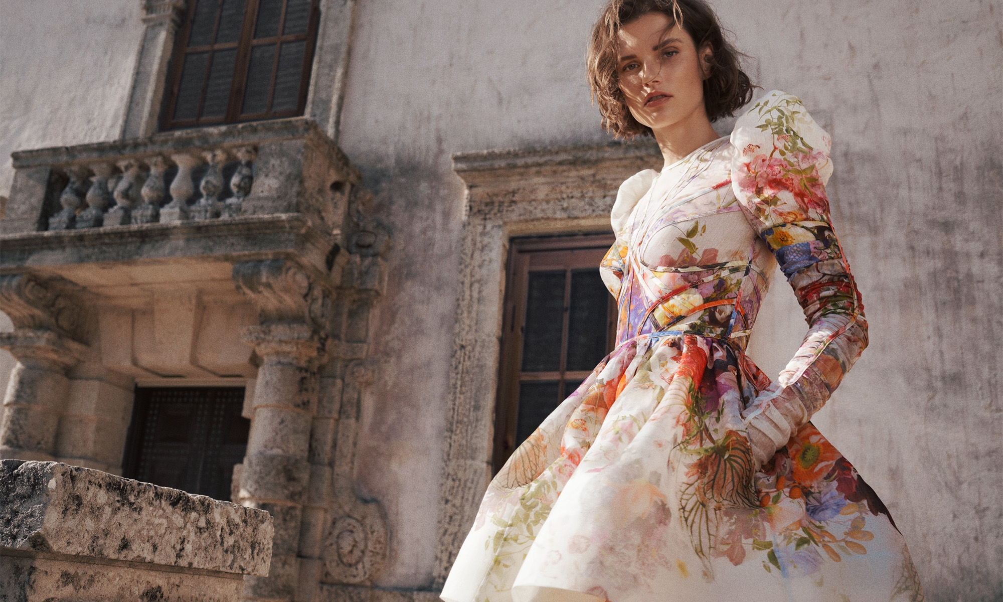 Adult female poses outdoors wearing a floral print dress with architectural background in medium shot, fashion editorial photography.