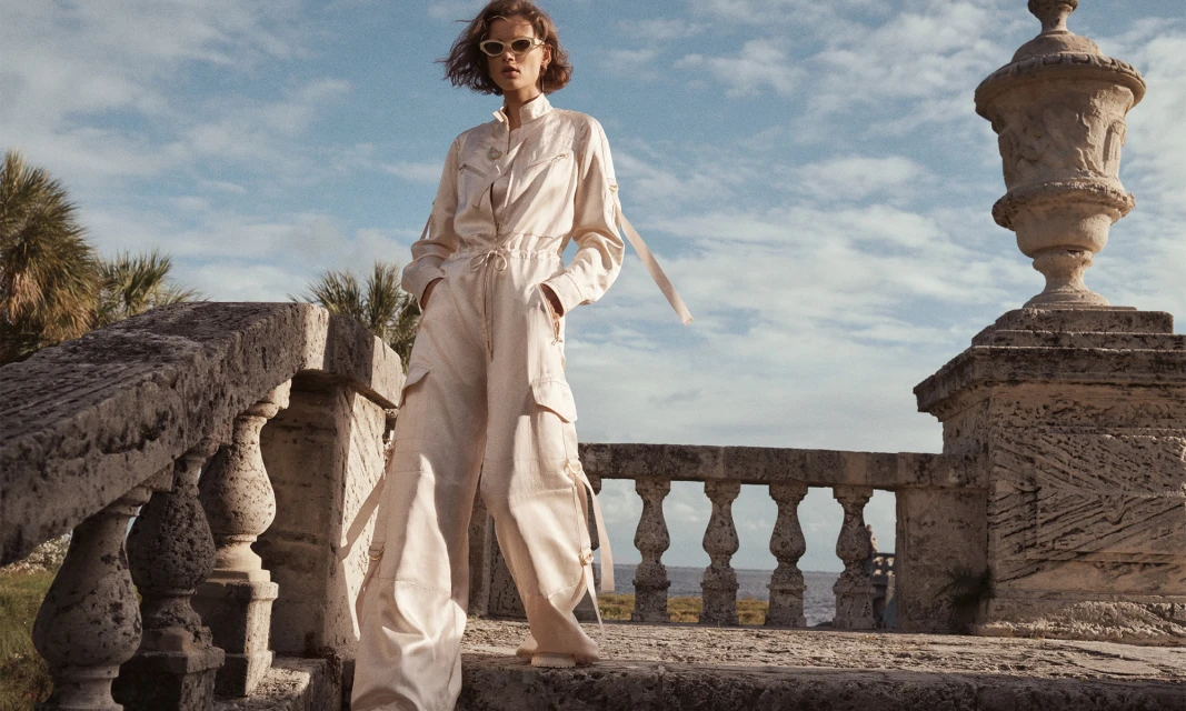 A low angle shot showcases a woman in a cream jumpsuit and sunglasses standing on a stone railing with ocean and sky in the background.